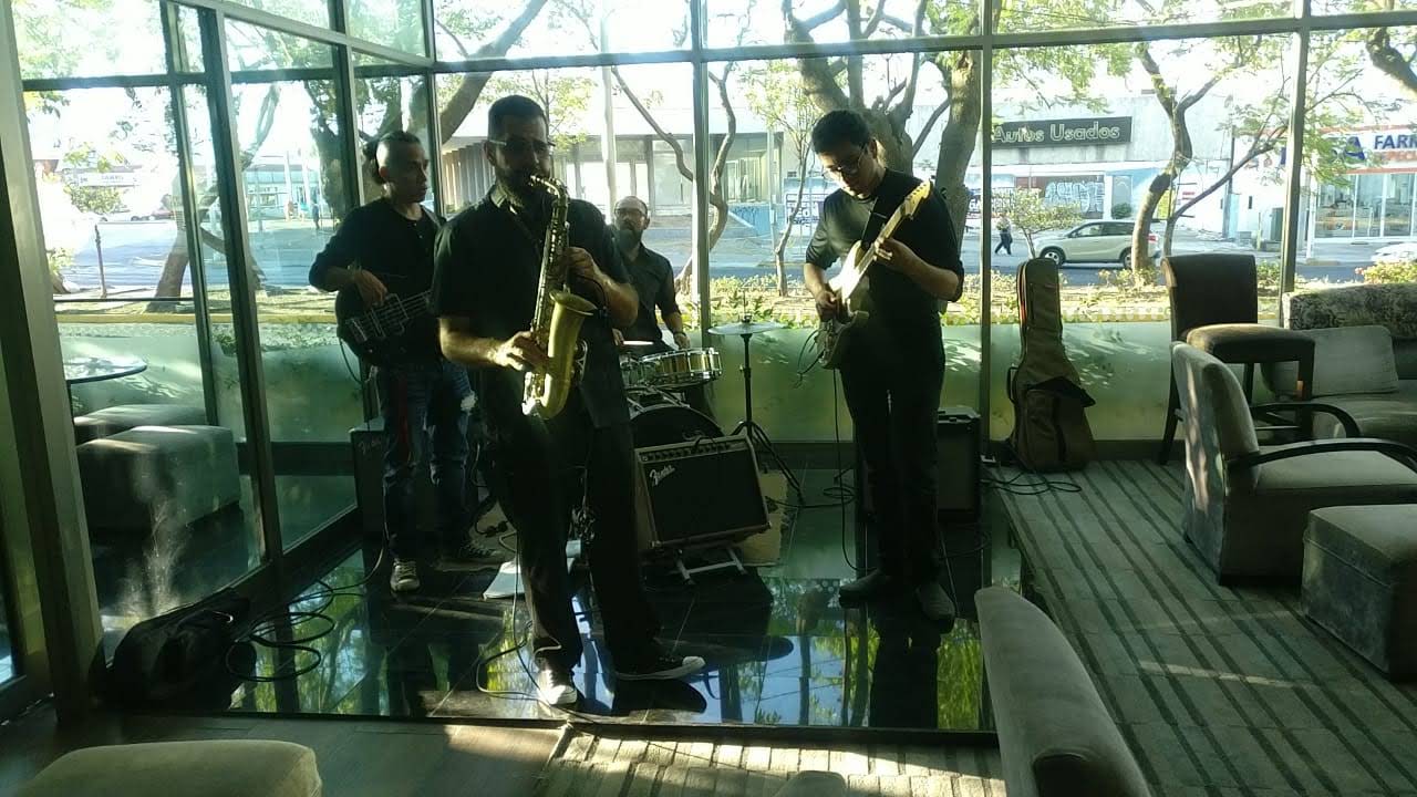 Guillermo toca guitarra eléctrica junto a un saxofonista y sección rítmica en el salón de un hotel.