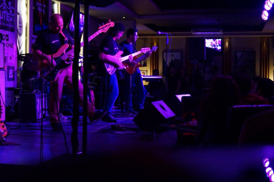 Guillermo toca guitarra eléctrica con una banda bajo luces violetas en un foro nocturno.